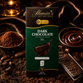 A box of Ahmia's 55% Sugar-Free Dark Chocolate chocolate bar is centered against a dark, richly styled background with chocolate sauce, cocoa powder, and chocolate curls.